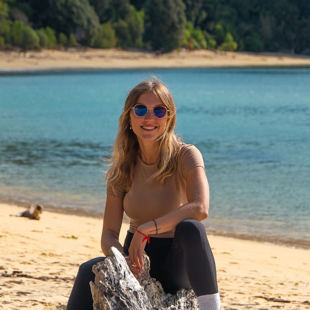 Jeune femme assise sur une plage équipée des Ushuaia sun devant un lac et une forêt